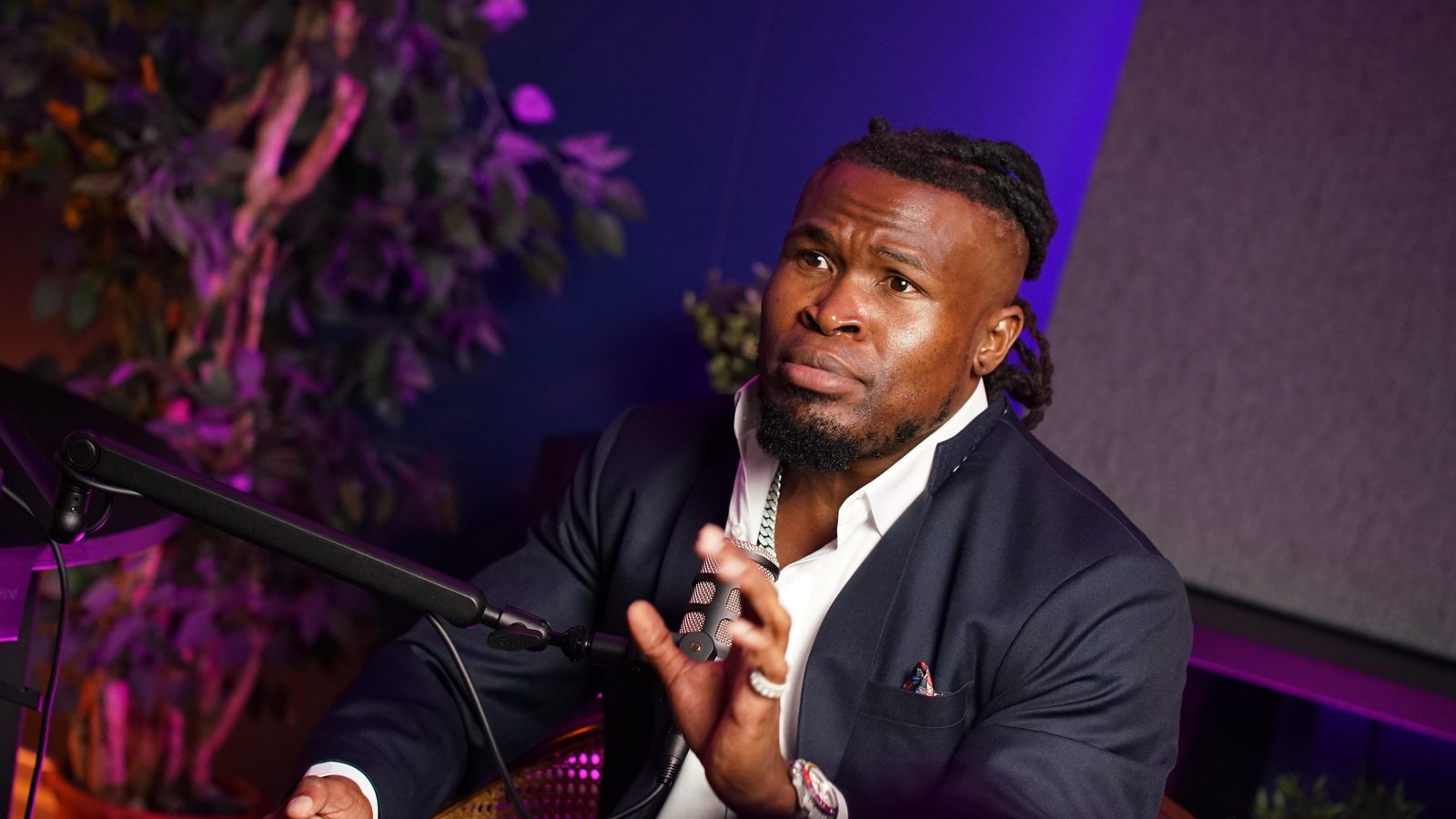 A guest in a navy suit talking with his hands at the New York set, plant and purple wall light behind him