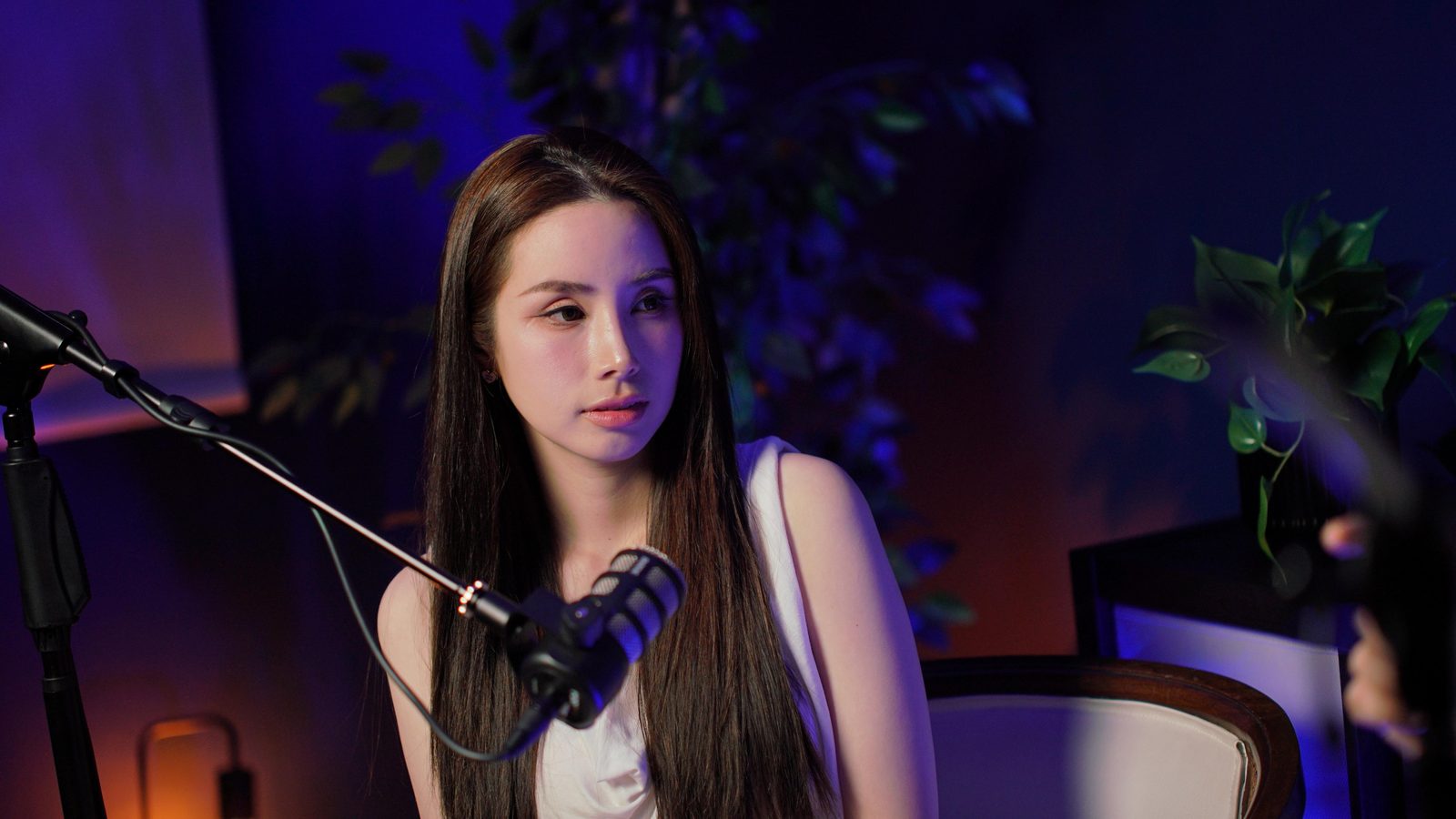 A guest in a white top at the New York set microphone, warm lamp glowing at the edge of frame