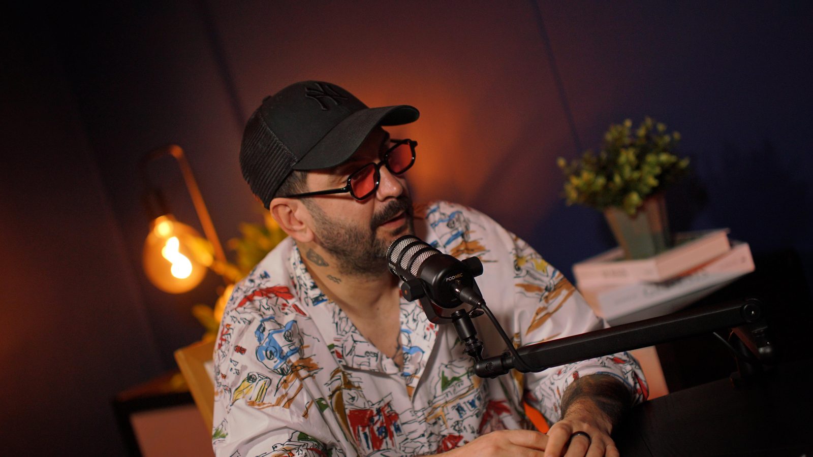 A guest in a cap and patterned shirt talking into the Paris set microphone, lamp and plant behind him