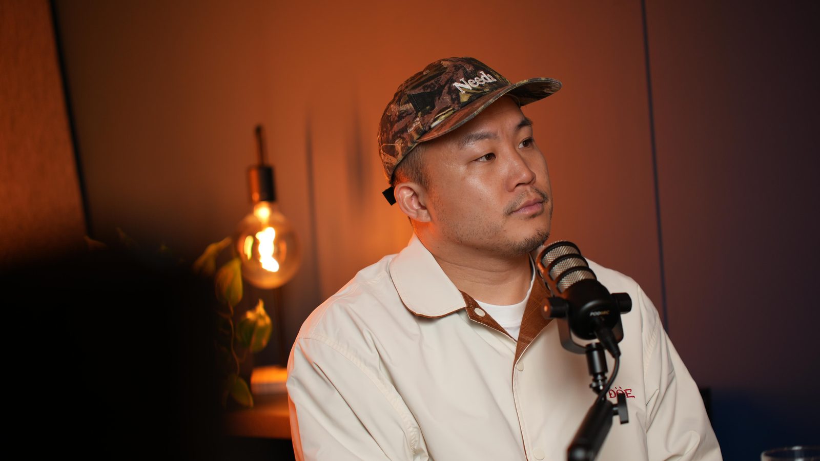 A guest in a cream jacket and cap listening at the Shanghai set, warm bulb and plant behind him