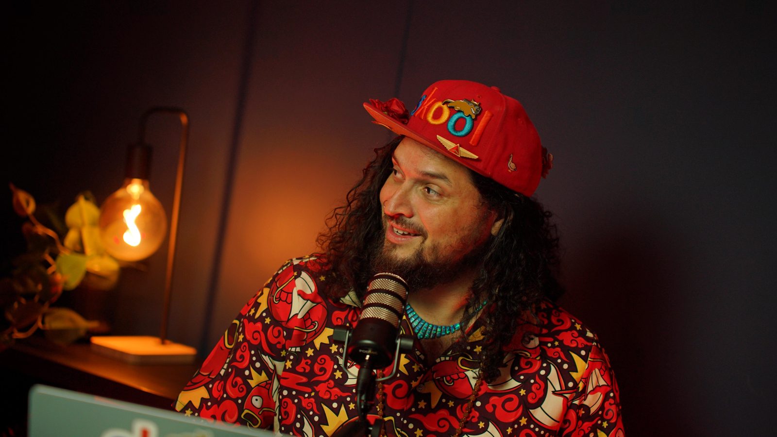 A guest in a bright patterned shirt talking at the Shanghai set, laptop and warm lamp in frame
