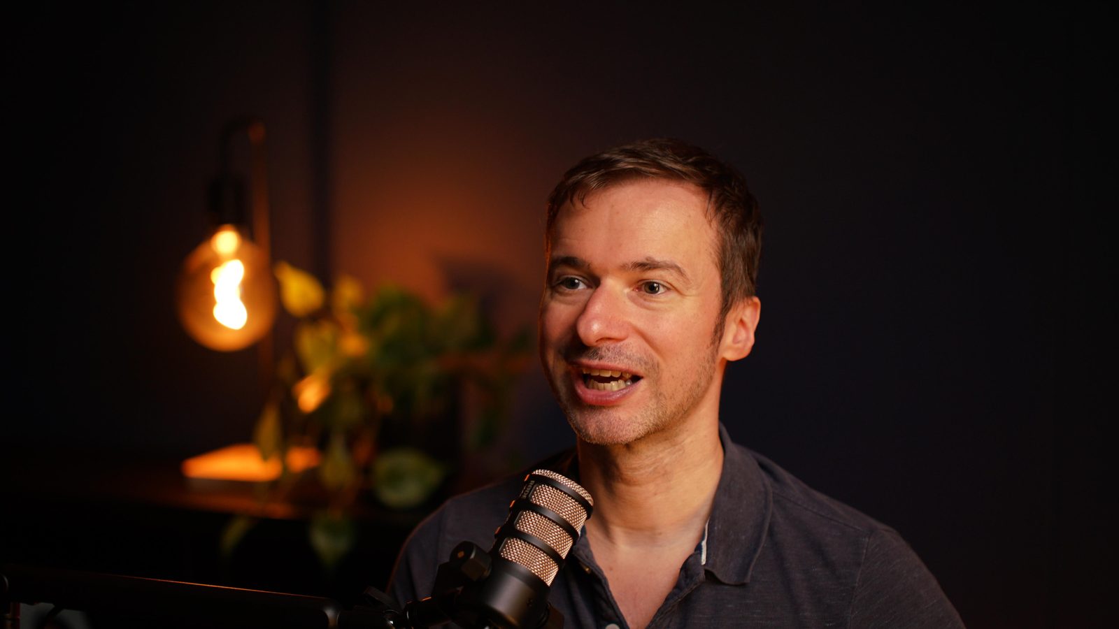 A guest in a grey polo mid-sentence at the Shanghai set microphone, warm bulb glowing behind