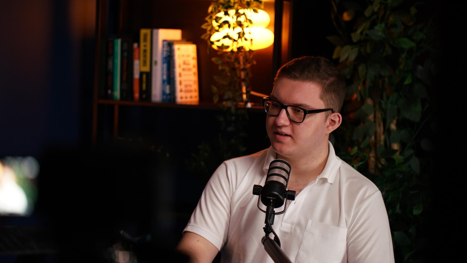 A guest in a white polo reading from a phone at the Shanghai set, lit bookshelf behind him