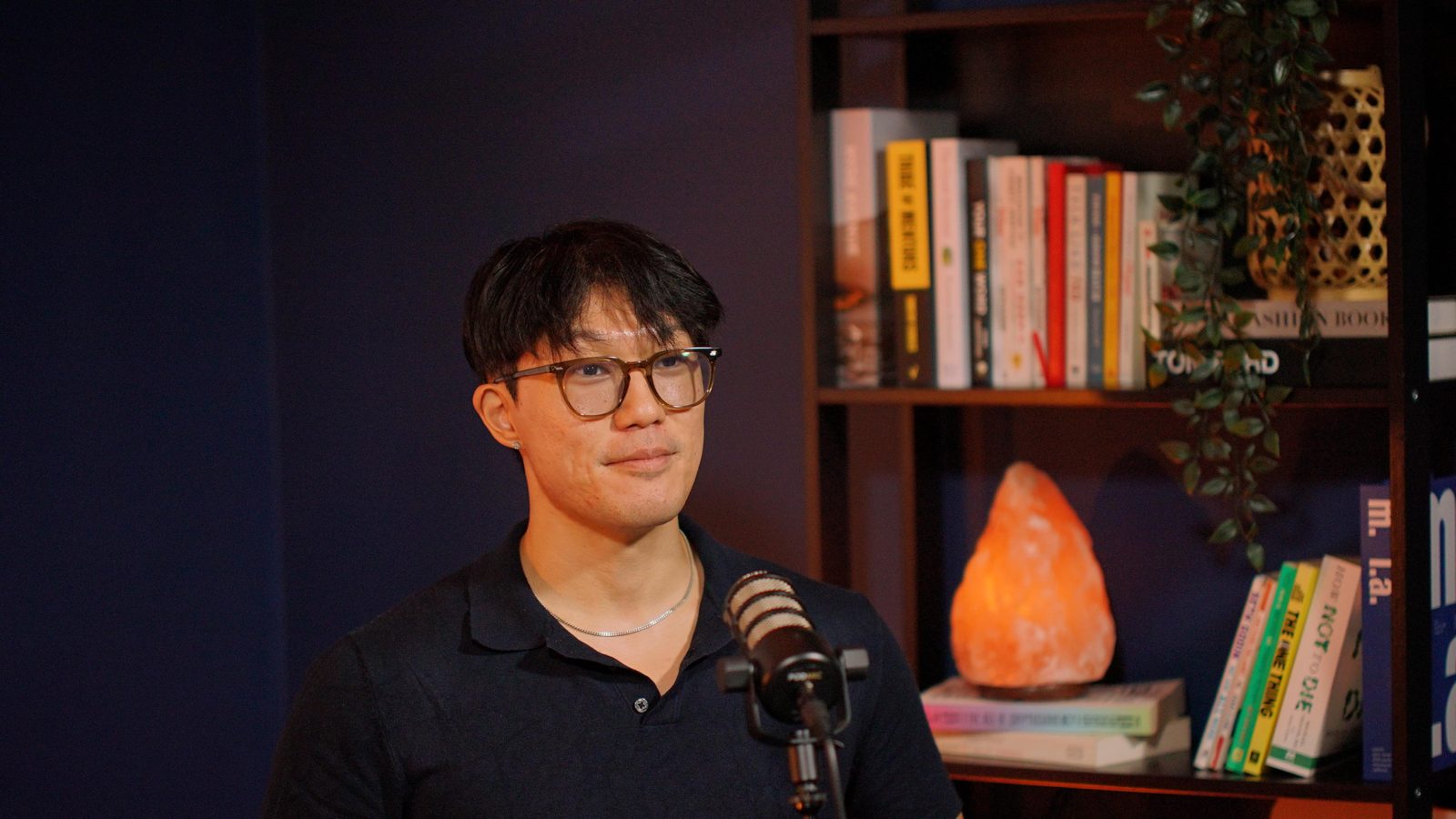 A guest in a navy polo at the Shanghai set, styled bookshelf and salt lamp filling the background