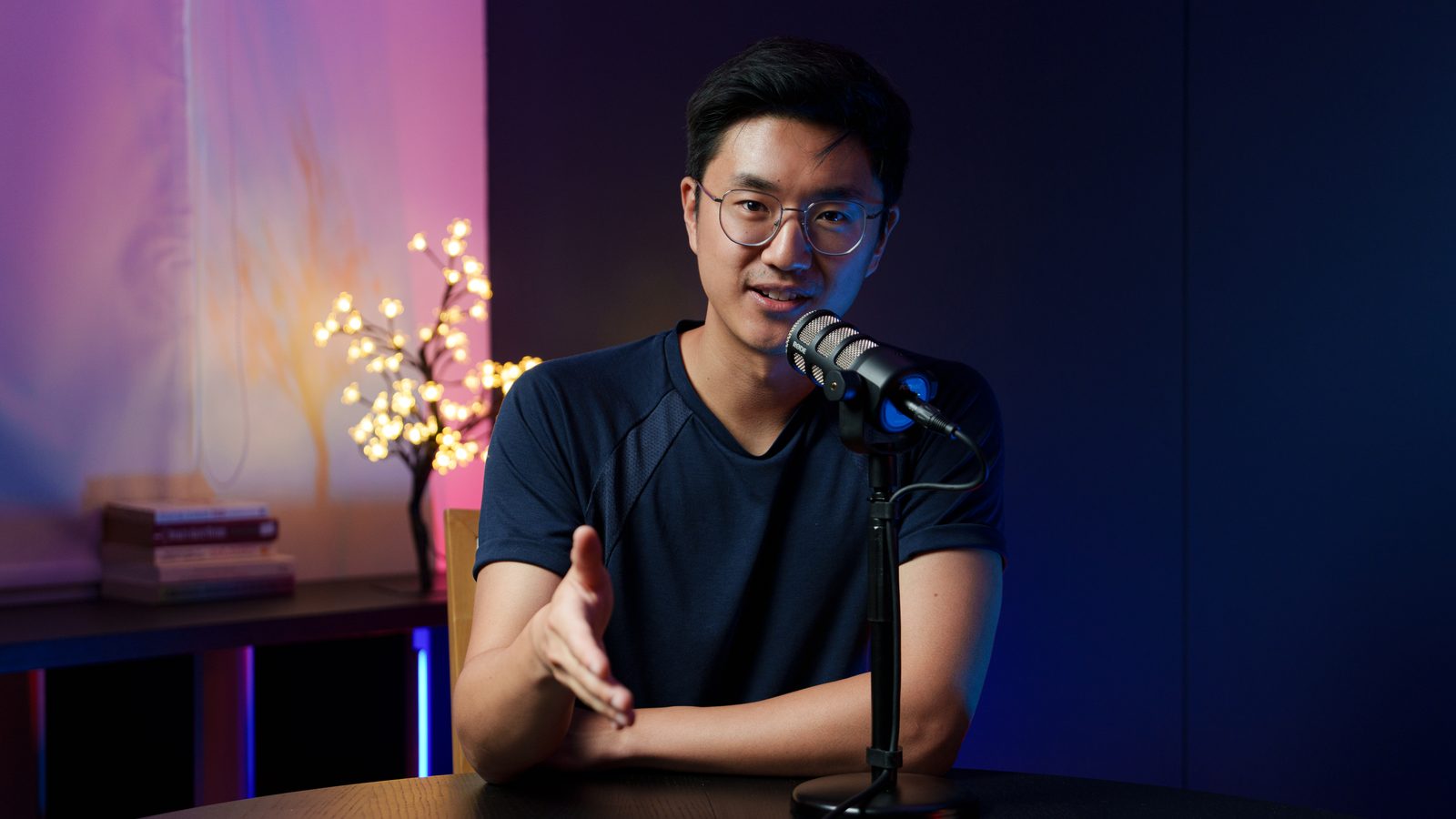 A guest in a navy t-shirt gesturing at the Shanghai set desk, lit branch and colour-washed wall behind